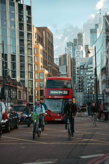 A busy urban street scene in Hackney with tall modern office buildings and commercial structures, featuring a red double-decker bus displaying 'Hackney Wick 26' on its front, surrounded by pedestrians, cyclists, and parked cars. The street is well-lit with natural daylight and shows clean surfaces with smooth asphalt, glass windows reflecting the sky, and contemporary architectural styles. This image represents an active city environment. For professional cleaning services, including surface cleaning and deep cleaning, visit Cleaner Hackney at cleanerhackney.co.uk for more information on affordable cleaners in Hackney.