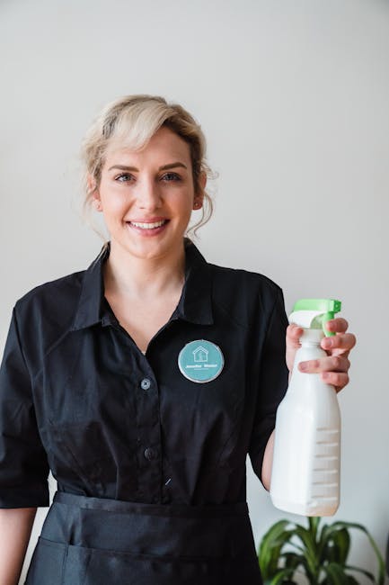 A woman with short blonde hair, dressed in a black shirt and apron, is standing in a bright room with white walls. She is smiling and holding a white spray bottle with a green nozzle, used for surface cleaning or sanitisation. In the background, there is a small potted plant placed on a surface, adding a touch of greenery to the scene. The lighting is natural and even, highlighting the clean and tidy environment. This image represents professional domestic or commercial cleaning services provided by Cleaner Hackney, specializing in surface cleaning, deep cleaning, and hygienic maintenance in residential and commercial settings within the Dalston E8 area.