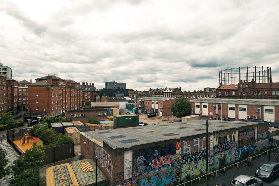 A scenic view of a narrow canal in Hackney, lined with lush green trees on both sides, some of which extend over the water. Along the right side, there is a paved walkway with a few pedestrians, and s