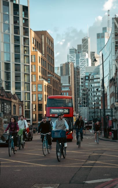 A busy city street scene in Hackney with modern glass and brick office buildings and residential structures. The street is filled with pedestrians and cyclists, including several people riding bikes, some wearing casual clothing. A red bus with route information 'Hackney Wick 26' is visible amidst the traffic. The street appears clean and well-maintained under partly cloudy skies, with natural daylight illuminating the area. This urban environment exemplifies a typical bustling London neighborhood, and Cleaner Hackney specializes in surface cleaning and hygienic maintenance for such commercial and residential spaces, ensuring a pristine and safe living and working environment.