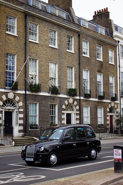 A three-story residential building with a traditional brick facade, featuring white-framed sash windows and black wrought iron balconies with potted plants on the second floor. The ground floor has arched white doorways and window frames. In front of the building, a black London taxi is parked on the street, which has clear road markings and a sidewalk with a black-and-white street sign. The scene is illuminated by natural daylight, highlighting the clean and well-maintained appearance of the building and surroundings. This image relates to cleaning services offered by Cleaner Hackney, emphasizing the importance of surface cleaning and maintenance in residential properties at the Mare Street end of tenancy cleaning guide in Hackney.