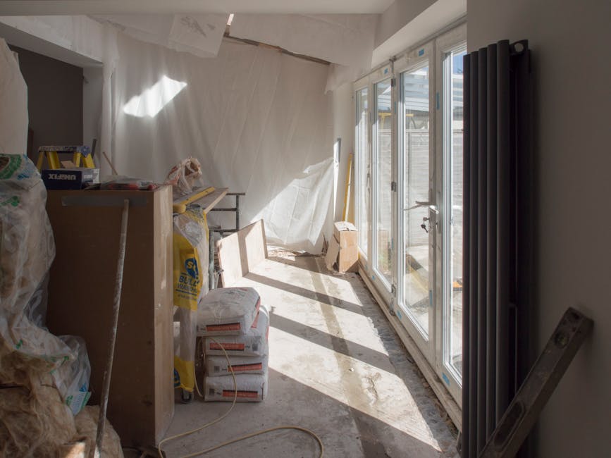 An interior room in a residential property under renovation, featuring a mixture of materials and surfaces. The foreground displays a wooden floor with visible dust and some stains, alongside a profes